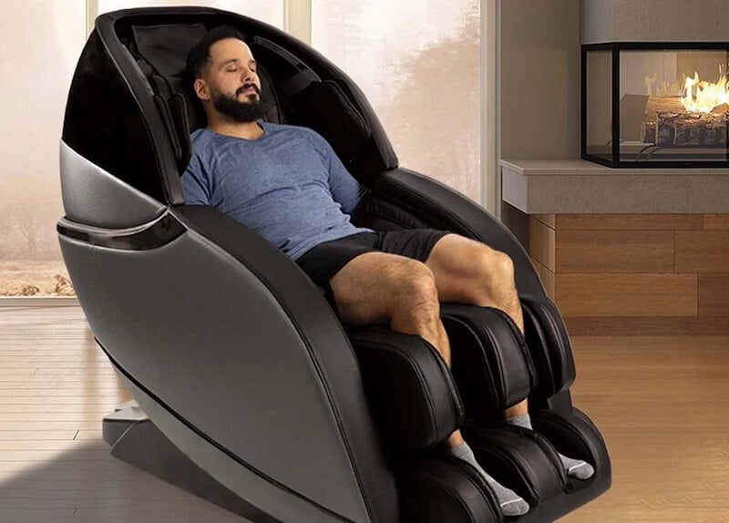 A relaxed man with a beard is reclining in a modern, black massage chair in a cozy room with a fireplace in the background. The man appears calm and content, eyes closed, enjoying the comfort of the chair