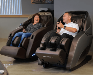 An image of two members of the Massage Chair Store Review Team testing massage chairs. The tester on the right reclines in a Brown Infinity Genesis Max 4D massage chair. 