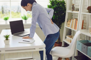 A woman in a blue shirt and jeans stands leaning forward at a white desk, touching her lower back in a gesture of discomfort. In front of her is an open laptop and a notebook, suggesting she may be taking a break from work due to back pain. The setting is a bright, home office with plants and bookshelves in the background.