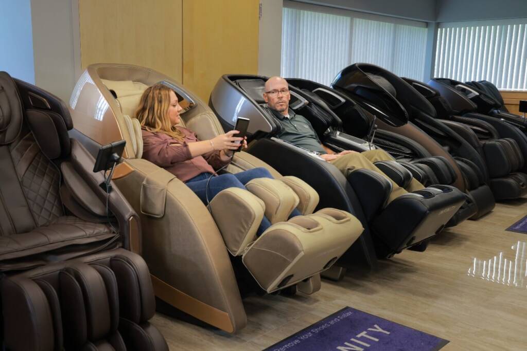 A member of the MCS review team testing the features of a brown Infinity Genesis Max massage chair, she is showing the controls to another team member sitting next to her in a black Infinity massage chair.