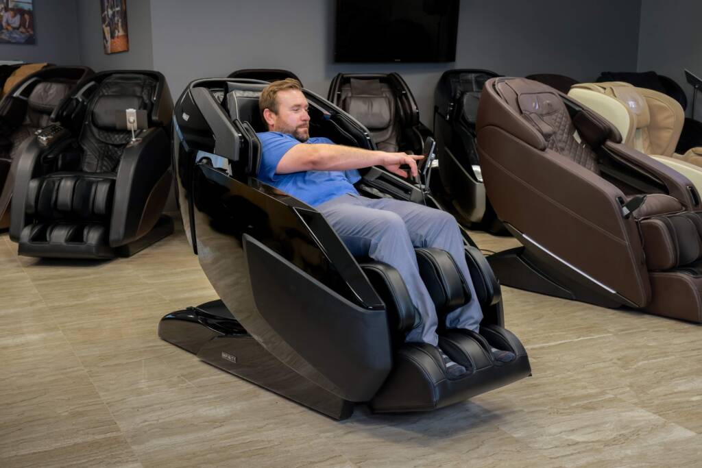 A member of the MCS review team sitting in a black Infinity Imperial Syner-D massage chair and using the touchpad controls. He is surrounded by other massage chairs within the MCS show room.