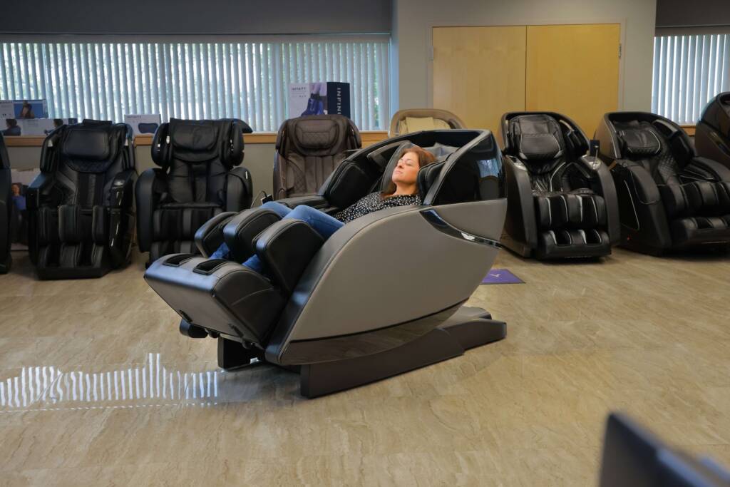 A member of the MCS review team reclining in a gray and black Kyota Kansha M878 massage chair. She is surrounded by other massage chairs within the MCS show room.