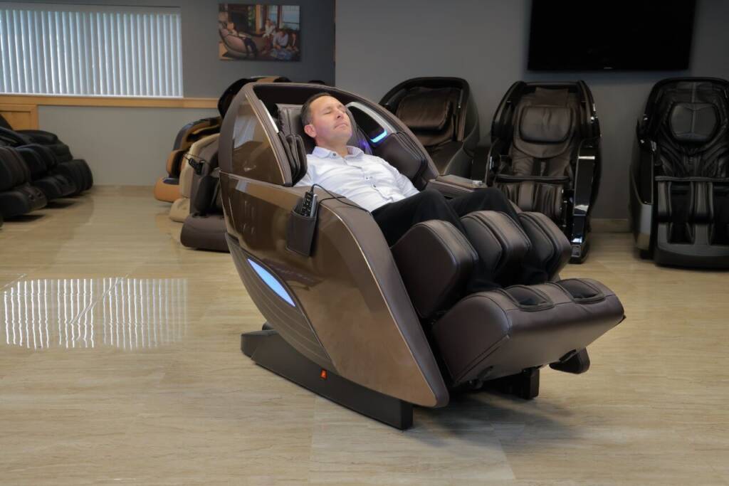 A member of the MCS review team reclining in a brown Kyota M989 Yutaka massage chair. He is surrounded by other massage chairs within the MCS show room.