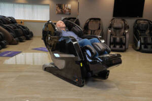 A member of the Massage Chair Store Review Team reclines in a black Kyota Yosei M868 4D massage chair, surrounded by other massage chairs in the MCS test facility.