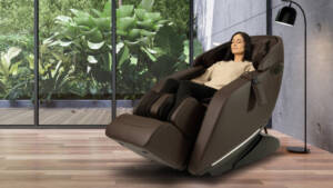 A woman is relaxing in a brown massage chair in an indoor patio with hardwood floors and floor-to-ceiling windows.