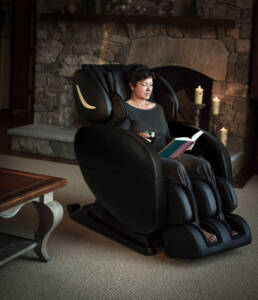 A woman is seated in a black massage chair in front of a stone fireplace, reading a book and enjoying a glass of wine.