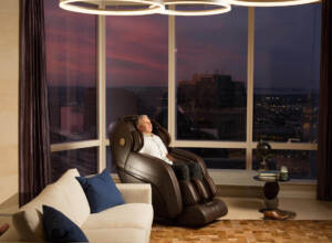 A man sits in a brown massage chair in a luxury high-rise apartment; the tall widows look out over the city at sunset.