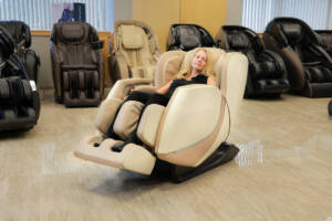 A member of the Massage Chair Store Review Team sits in a cream-colored Kyota E330 Kofuko massage chair, surrounded by other massage chairs in the MCS test facility.
