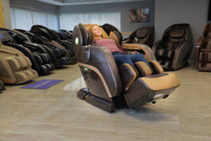 A member of the Massage Chair Store Review Team reclines in a brown Kyota M888 Kokoro massage chair, surrounded by other massage chairs in the MCS test facility.