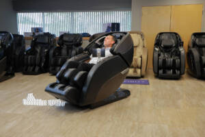 A member of the Massage Chair Store Review Team reclines in a black Kyota Nokori M980 massage chair within the MCS test facility. The massage chair's control panel is flipped upright and facing the user.