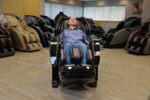 A member of the Massage Chair Store Review Team reclines in a black Kyota Yosei M868 4D massage chair, surrounded by other massage chairs in the MCS test facility.