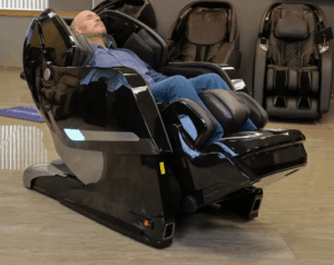 A member of the Massage Chair Store Review Team reclines in a black Kyota Yosei M868 4D massage chair, surrounded by other massage chairs in the MCS test facility.